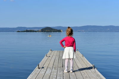 Rear view of girl with hand on hip standing on pier against sky