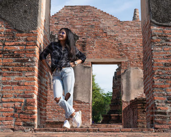 Full length of woman sitting on brick wall of building
