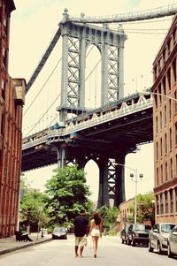 Full length rear view of friends walking on city street against manhattan bridge