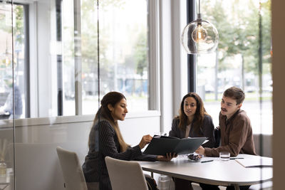 Female real estate agent showing floor plan to young customers while sitting at desk in office