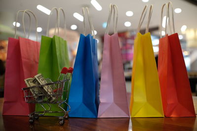 Close-up of colorful shopping bags with paper currencies in miniature shopping cart on wooden table