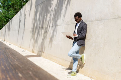 Focused african american male executive manager standing against concrete wall in the street with laptop