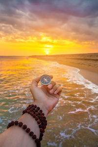 Cropped hand holding navigational compass at beach against sky during sunset