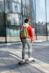 Hipster in red jacket, sunglasses, jeans, white sneakers and with green backpack riding on longboard