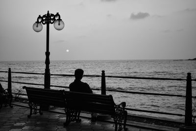 Rear view of man sitting on bench by sea against sky at sunset