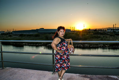 Portrait of woman standing by railing against river and sky during sunset