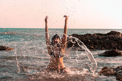 Woman splashing water in sea against clear sky at sunset