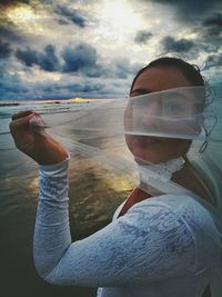 Portrait of playful bride holding veil at beach against cloudy sky during sunset