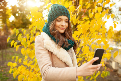 Young woman in warm clothing taking selfie through smart phone while standing against autumn tree