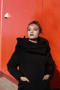 Fashionable teenage girl wearing black warm clothing standing against red wall during sunny day