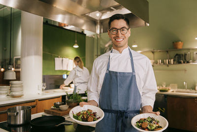 Portrait of confident male chef showing food while standing in commercial kitchen