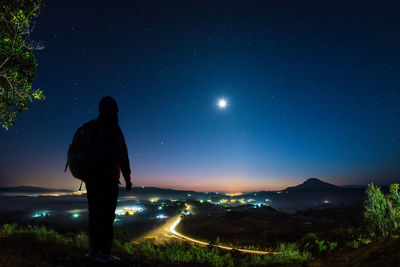 Rear view of man standing on illuminated street against sky at night