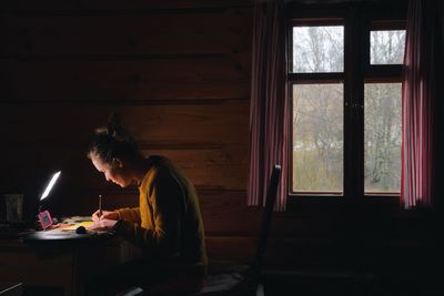 Side view of woman writing on table by window in log cabin