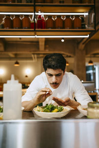 Focused male chef sprinkling salt on salad while bending in commercial kitchen