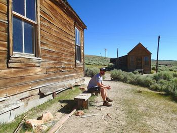 Full length side view of man sitting on bench by house during sunny day