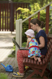 Mother using laptop while sitting with daughter on bench in yard