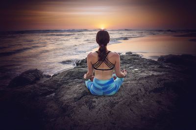 Rear view of woman sitting on rock at beach during sunset