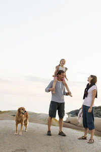 Mother looking at girl enjoying sitting on shoulder of father standing against sky