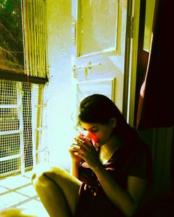 Woman drinking coffee in cup while sitting by window at home