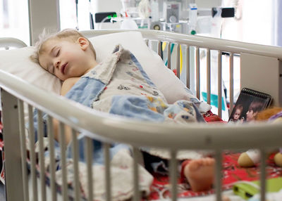 Toddler sleeping in hospital crib with toys and family photos.