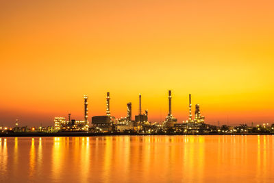 Illuminated factory by river against sky during sunset