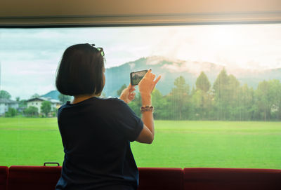 Rear view of woman photographing through mobile phone from train window