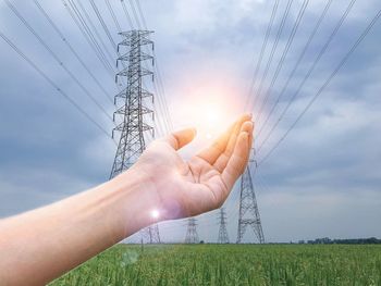 Cropped hand with electricity pylon in background against cloudy sky