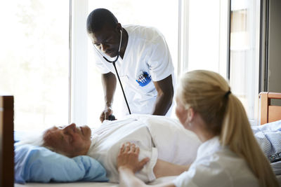 Female nurse consoling senior man while male colleague examining with stethoscope in hospital ward