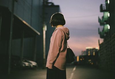 Side view of woman standing on city street against sky at dusk