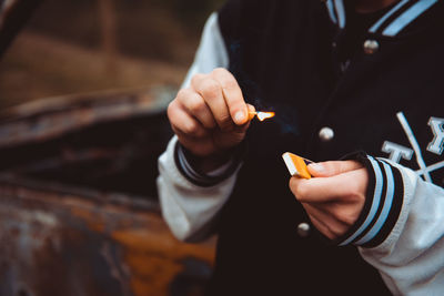 Unrecognizable person in casual outfit igniting match while standing on blurred background of old rusty car person