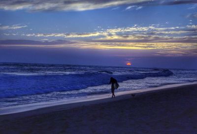 Silhouette person holding surfboard while walking at sea shore during sunset