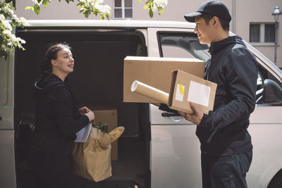 Delivery woman talking to smiling colleague while holding package near truck