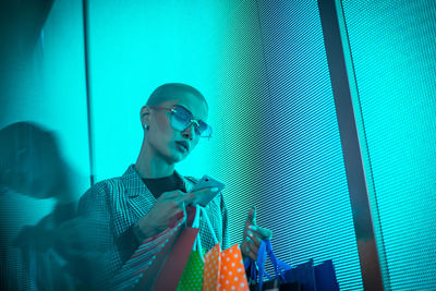 Low angle view of lesbian woman using smart phone holding shopping bags standing against abstract backgrounds