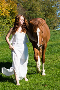 Young woman with horse walking on grassy field against trees during sunny day