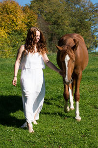 Young woman with horse walking on grassy field against trees during sunny day