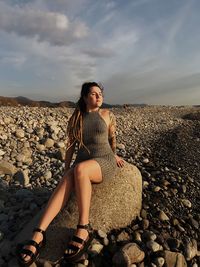 Young woman sitting on pebble stones at beach against sky