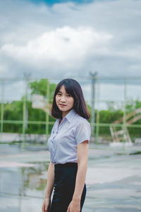 Portrait of smiling young woman standing on wet road against cloudy sky during rainy season