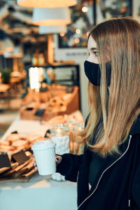 Woman holding coffee cup, standing in queue in a coffee shop. person wearing face mask