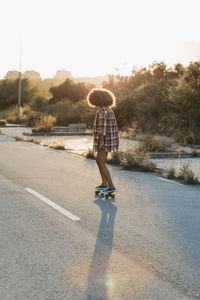 Back view of female hipster with afro hairstyle riding penny board along empty road during sunset