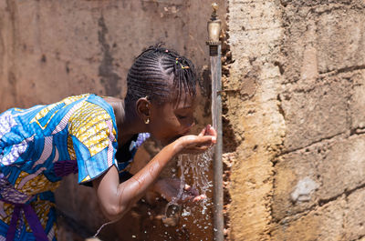 Girl drinking water from faucet against wall