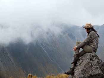 Woman sitting on rock while looking at rocky mountains against sky