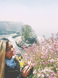 Young woman smelling flowers while standing on mountain during sunny day