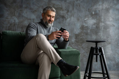 Senior handsome man sitting on green couch in loft living room, working from home on tablet computer