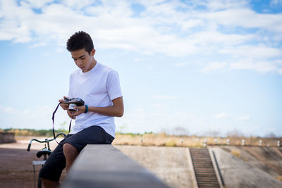 Teenage boy using camera while sitting on table against sky