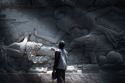 Rear view of a man standing facing a cliff with a relief sculpture of the ramayana story