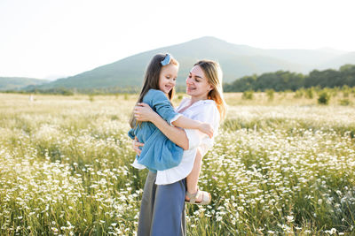 Cute young woman holding hands and play with child girl in chamomile field with blooming flower