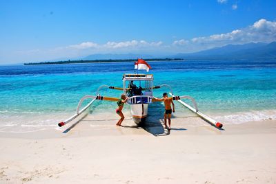 Boys standing by boat at beach against blue sky during sunny day