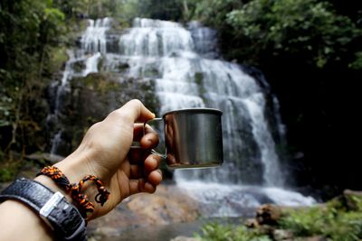 Cropped hand holding cup against waterfall in forest