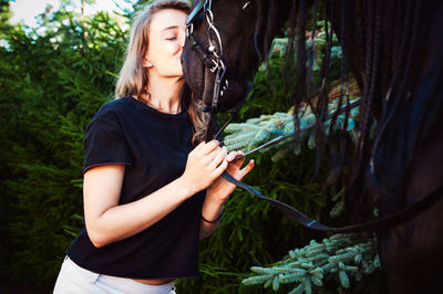 Young woman kissing horse while standing by trees