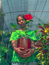 Low angle portrait of young woman holding potted plant while standing in greenhouse